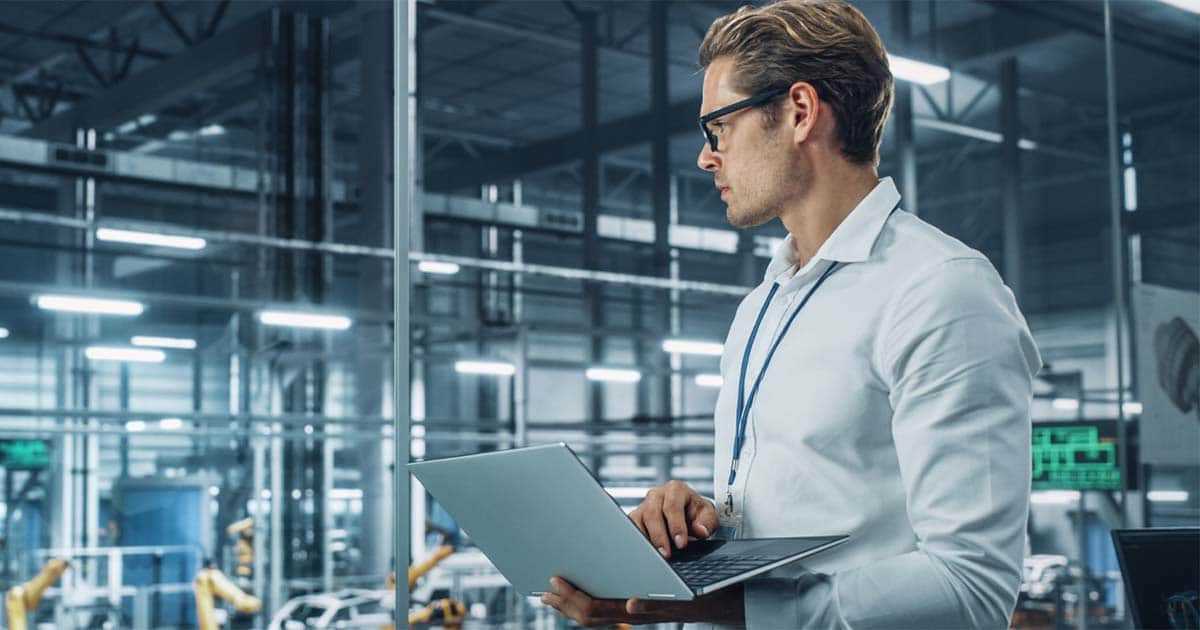 Eine Person in Businesskleidung steht mit Laptop vor einer Maschine