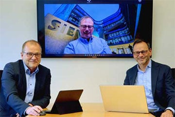 Zwei Männer sitzen am Tisch und arbeiten an ihren Computern, während ein Fernsehbildschirm im Hintergrund läuft